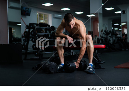 Muscular shirtless man sitting on a bench and lifting a dumbbell.  110098521