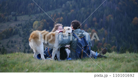 Caucasian couple of hikers sits on the hill, pets and plays with local dog 110101692