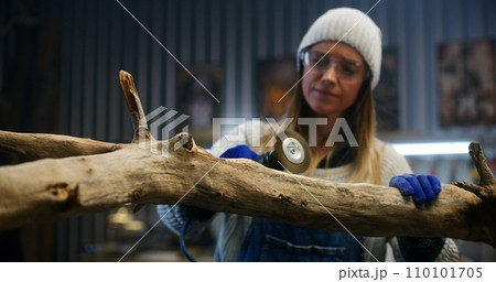 Female carpenter in goggles grinds tree branch using grinding machine Female carpenter in goggles grinds tree branch using grinding machine 110101705