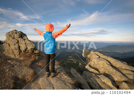 Young child boy hiker standing with raised hands in mountains enjoying view of amazing mountain landscape at sunset. 110102494