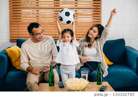 A jubilant family, with popcorn and a ball, raises their arms, cheering while watching a football match at home. Their togetherness and excitement reflect the joy of winning. 110102785