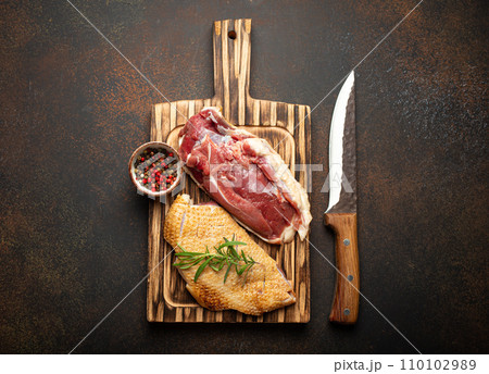 Two raw uncooked duck breast fillets with skin, seasoned with salt, pepper, rosemary top view on wooden cutting board with knife, dark brown concrete rustic background Two raw uncooked duck breast fillets with skin, seasoned with salt, pepper, rosemary top view on wooden cutting board with knife, dark brown concrete rustic background 110102989