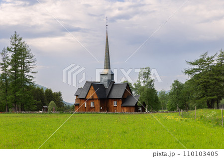 Vibrant green field and wooden Kvikne Kirke, surrounded by lush trees under cloudy sky 110103405