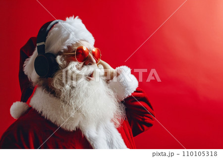 AI-generated content. Close-up portrait of Santa Claus in trendy sunglasses wearing wireless headphones. Advanced Santa listens to his favorite music. Red background with copy space. 110105318