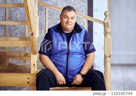 Sly smile on face of large European man, 47 years old, sitting on steps of stairs leading to second floor of his house. 110105744