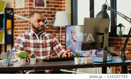 Overworked teleworker carefully typing data on computer in warm cozy living room apartment listening to tv background noise. Focused tired freelancer remotely working stressed by deadlines 110107793