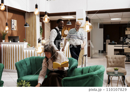Friendly african american bellhop assists senior man in hotel lobby with luggage to the reserved room. Elderly female visitor reads while waiting for accomodation in cosy lounge area with armchairs. 110107829