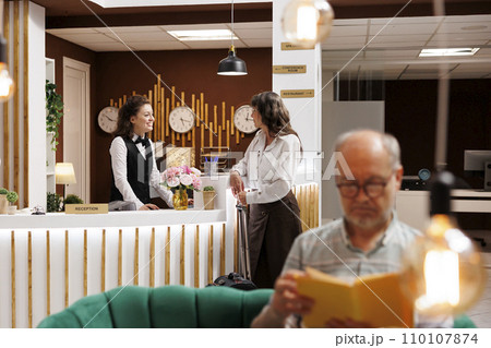 Elderly female guest with luggage speaking to young receptionist at front desk of luxury resort. Old man with a novel relaxes in hotel lobby while senior woman does booking procedure at counter. 110107874