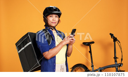 Portrait of courier waiting, looking in phone app for next customer order location. BIPOC woman delivering food with bycicle using thermic bag, isolated over studio background 110107875