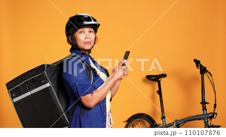 Courier delivering food, texting customer to pick up order. Asian woman in a rush, upset with client taking long time to get purchased meal, isolated over orange studio background 110107876