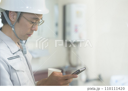 工場や現場の仕事で休憩中にスマホを見るヘルメットを被りメガネをかけた男性作業員 110111420