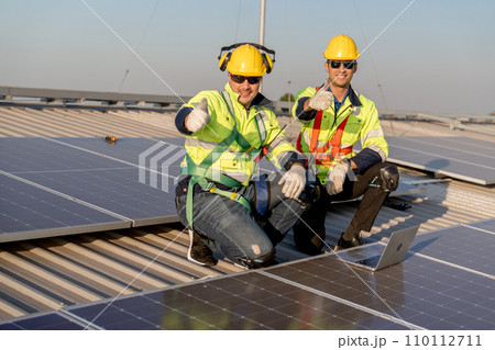Two industrial technician worker sit near solar cell panels on rooftop of factory building and show thumbs up to camera with smiling. Green and sustainable energy concept. 110112711