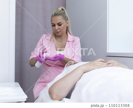 Esthetician Holds White Bottle with Peeling or Cleansing Foam, Cream above Beautiful Young Caucasian Female Patient. Beauty Procedure, Moisturizing Face Skin, Facial Massage. Horizontal Esthetician Holds White Bottle with Peeling or Cleansing Foam, Cream above Beautiful Young Caucasian Female Patient. Beauty Procedure, Moisturizing Face Skin, Facial Massage. Horizontal 110113308