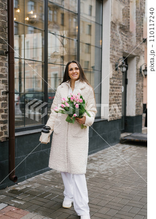 Young stylish woman with a takeaway coffee and a bouquet of tulips walking on a city street in springtime 110117240