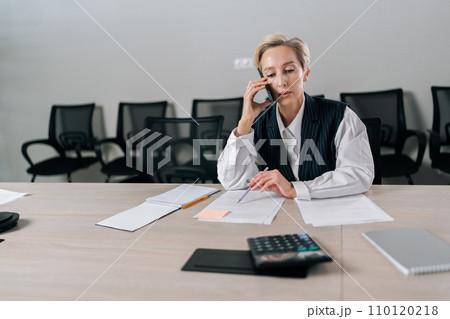 Portrait of middle-aged businesswoman making business call sitting on office desk, working with document. Front view of gray-haired female manager talking at smartphone with client discussing contract 110120218