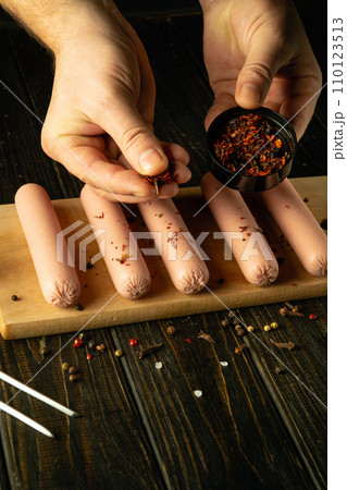 The cook adds spices to raw meat sausages before grilling. Concept of cooking fried Munich sausages on kitchen table by chef hands 110123513
