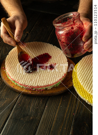 The chef's hand holds a spoon with jam while preparing a cake with condensed milk. Concept of preparing pastry dessert on restaurant kitchen table 110123514