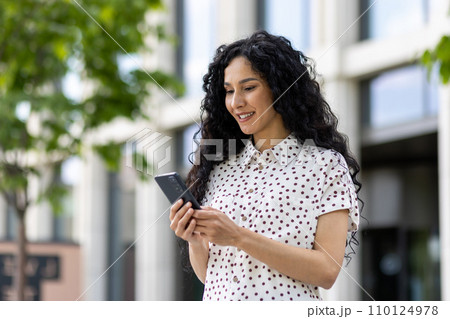 A young beautiful Latin American woman with curly hair walks through the city with a phone in her hands, a woman uses an application on a smartphone, browses Internet pages and social networks. A young beautiful Latin American woman with curly hair walks through the city with a phone in her hands, a woman uses an application on a smartphone, browses Internet pages and social networks. 110124978