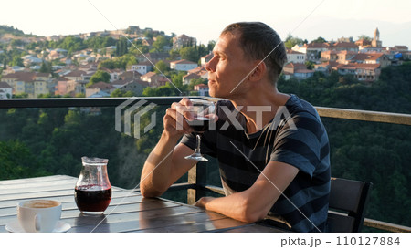 Thoughtful male drinking red wine while sitting at table of cafe or restaurant by scenic view of European town 110127884
