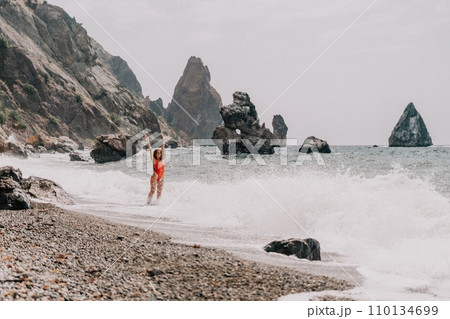Woman summer travel sea. Happy tourist in red bikini enjoy taking picture outdoors for memories. Woman traveler posing on beach at sea surrounded by volcanic mountains, sharing travel adventure joy 110134699