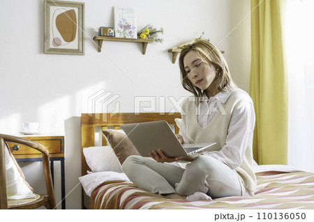 自宅のベッドの上に座ってノートパソコンを操作する茶髪の若い女性 110136050