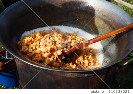 Stirring fat in a large vat on firewood. Pork rinds rendered, fried in fat, baked, roasted to produce pork cracklings or scratchings. Toasted brown cracklings. Large cast iron vat or cauldron. 110138021
