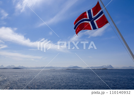 Vivid Norwegian flag billows against a summer sky, with snowy mountains 110140932