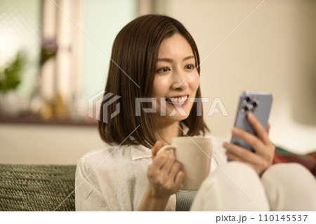 ソファーでくつろいでコーヒーを飲みながらスマホを見るミドル女性 110145537