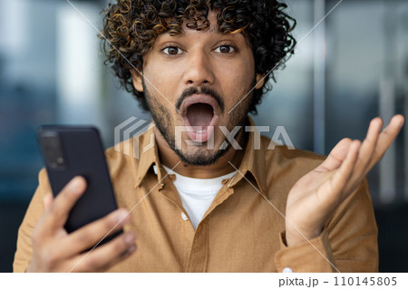 Close-up portrait of a young Indian man looking shocked and surprised at the camera while holding a mobile phone and spreading his hands. Close-up portrait of a young Indian man looking shocked and surprised at the camera while holding a mobile phone and spreading his hands. 110145805