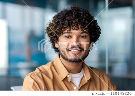 Close-up portrait of a young smiling Indian man sitting and working in the office, looking confidently at the camera. Close-up portrait of a young smiling Indian man sitting and working in the office, looking confidently at the camera. 110145820