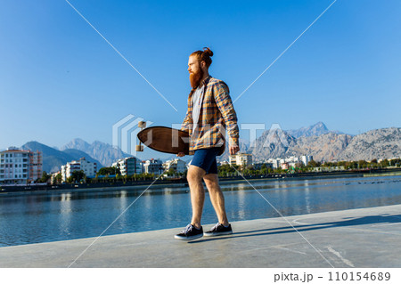 handsome redhaired man with long beard skateboarding near river at sunny summer day handsome redhaired man with long beard skateboarding near river at sunny summer day 110154689