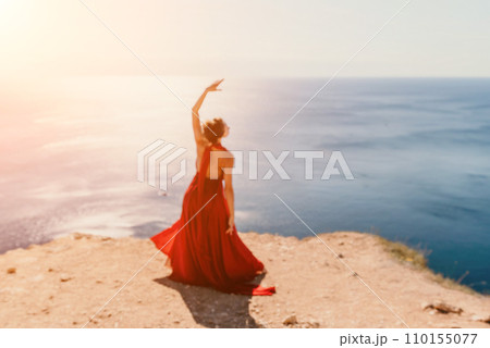 Woman in red dress on sea. Side view a Young beautiful sensual woman in a red long dress posing on a rock high above the sea on sunset. Girl on the nature on blue sky background. Fashion photo. 110155077