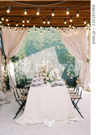 Laid table with bouquets of flowers stands on the terrace of an old house under a wooden canopy with curtains 110165868