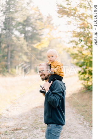 Smiling dad with a little boy on his shoulders stands half-turned in a clearing in the forest 110165908
