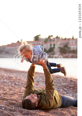 Dad is lying on the beach holding a little girl above him in outstretched arms 110165934