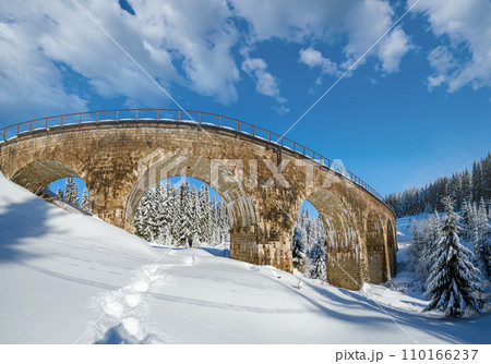 Stone viaduct (arch bridge) on railway through mountain snowy fir forest. Snow drifts  on wayside and hoarfrost on trees and electric line wires. 110166237