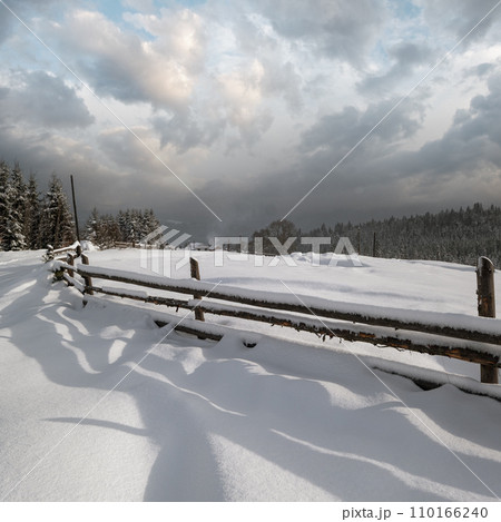 Picturesque waved shadows on snow from wood fence. Alpine mountain winter hamlet outskirts, snowy path, fir forest on far misty and cloudy hills. 110166240