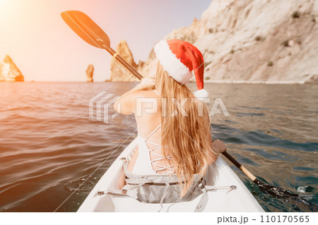 Woman in kayak back view. Happy young woman in Santa hat floating in kayak on calm sea. Summer holiday vacation and cheerful female people relaxing having fun on the boat. 110170565