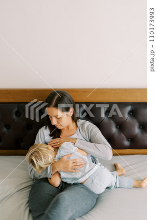 Mom breastfeeds a little girl hugging her in her arms while sitting on the bed 110173993