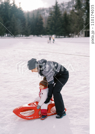 Mom puts a little girl on a sled on a snowy lawn 110174035