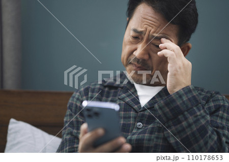 スマホを見ながら顔をしかめるパジャマ姿の50代の男性 スマホを見ながら顔をしかめるパジャマ姿の50代の男性 110178653