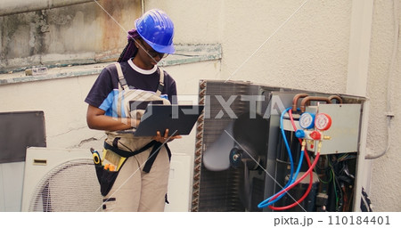 Qualified african american electrician working on outside hvac system, holding laptop. Licensed serviceman optimizing external air conditioner performance, ensuring it operates at maximum capacity 110184401