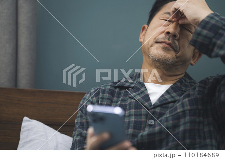 スマホを見ながら眉間を押さえるパジャマ姿の50代の男性 スマホを見ながら眉間を押さえるパジャマ姿の50代の男性 110184689