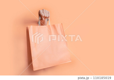 Female hand holding peach fuzz color paper bag trough the hole in paper backdrop 110185019