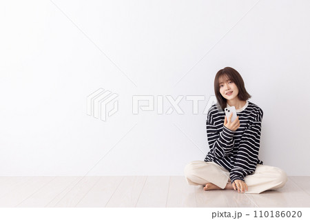 床に座りスマートフォンを使う若い女性 110186020