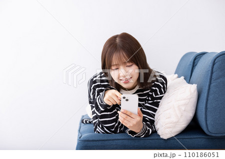 ソファーでスマホを寝そべりながら使う女性 110186051