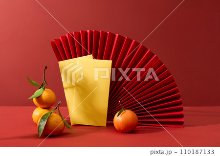 A large handmade paper fan featured over red background with envelopes and tangerines. The Chinese Lunar New Year is associated with many folk legends 110187133