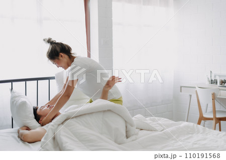 Funny couple wife try to wake lazy husband up in white bedroom. 110191568