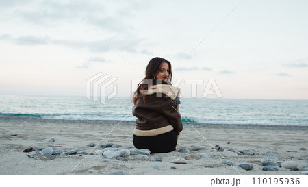 Girl seated on the beach near the ocean in overcast winter day Girl seated on the beach near the ocean in overcast winter day 110195936