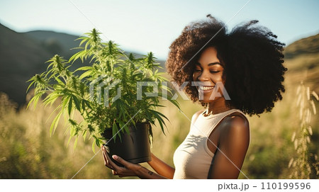 A woman holds a pot of flowering cannabis against a green field. 110199596